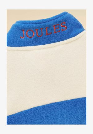 Zila un krēmkrāsas flīsa apģērba apkakle ar sarkanu izšūtu "JOULES" tekstu aizmugurē.