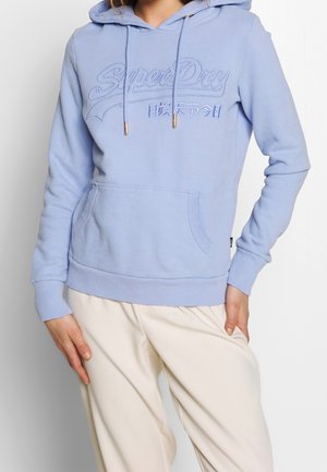 Kobieta ubrana w jasnoniebieską bluzę SuperDry z kieszenią kangurzą i kremowe spodnie, z ręką opartą na biodrze i udzie.