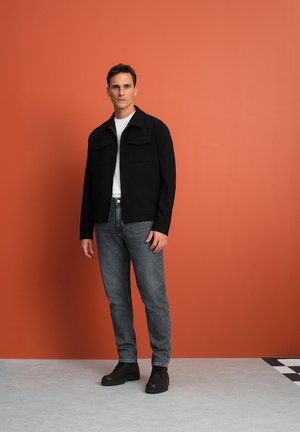 Czarna kurtka z dwiema kieszeniami na klatce piersiowej, biała koszulka pod spodem, szare dżinsy i czarne botki, na pomarańczowym tle.