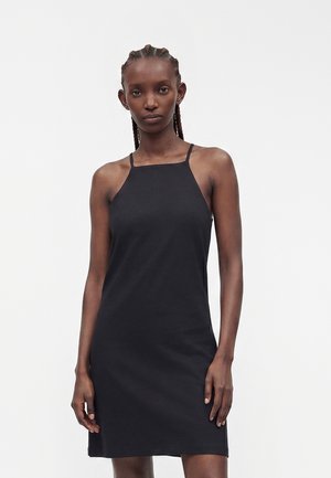 Femme aux cheveux tressés portant une robe noire sans manches arrivant aux genoux avec de fines bretelles sur un fond clair uni.