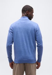 Άνδρας που κοιτάζει προς τα πίσω, φοράει ανοιχτό μπλε πουλόβερ με ζιβάγκο και μπέζ παντελόνι, με φόντο ένα λευκό απλό τοίχο.