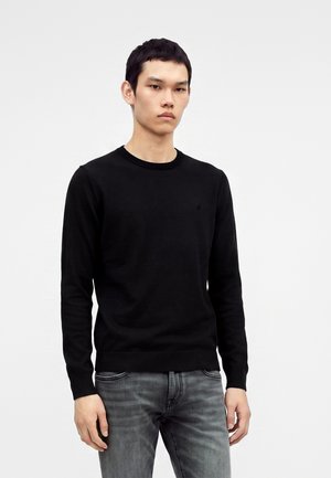 Joven con el pelo corto y oscuro, vestido con un suéter negro de manga larga y vaqueros grises, de pie frente a un fondo liso y claro.