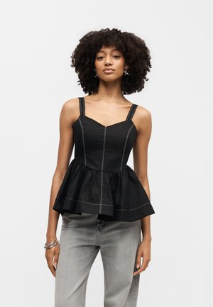 Γυναίκα με μαύρο αμάνικο peplum τοπ με λευκές ραφές και ανοιχτό γκρι τζιν, στέκεται μπροστά σε λευκό φόντο.