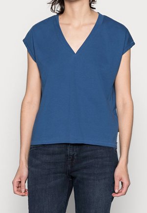 Person iført en kortærmet blå V-hals T-shirt og mørke jeans, stående mod en ensfarvet lys baggrund.