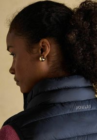 Jūras zila pūkainā veste ar quiltotu tekstūru un "JOULES" zīmolu. Apaļas zelta auskari redzami, modelis attēlots profila skatījumā uz neitrāla fona.