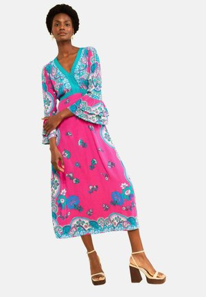 Femme portant une longue robe rose et turquoise à motifs avec des manches évasées et des sandales à talons hauts beiges, debout sur fond blanc.