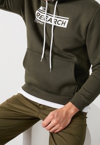 Sudadera oliva con bolsillo frontal, cordones blancos y un gráfico blanco que dice "RESEARCH". Combinada con pantalones oliva.