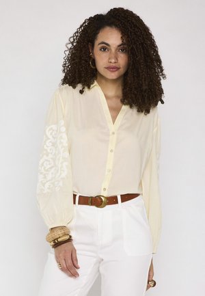 Femme aux cheveux bouclés portant un chemisier crème boutonné avec des manches brodées, un pantalon blanc, une ceinture marron et plusieurs bracelets au poignet gauche.