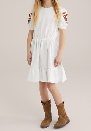 Ung person stående iført en hvid tekstureret kjole med broderede blomsterærmer og brune ruskind cowboystøvler.