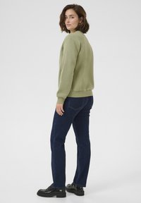 Felpa verde oliva con orli e polsini a coste, abbinata a jeans dritti blu scuro e mocassini neri con zeppa. Modella di profilo.