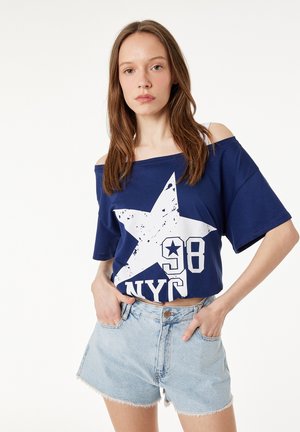 Młoda kobieta o brązowych włosach, ubrana w granatowy t-shirt z grafiką gwiazdy NYC odsłaniający ramiona oraz jasne jeansowe szorty z wysokim stanem, stoi z rękami w kieszeniach.