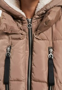 Taupe pufferjakke med frontglidelås og to vertikale glidelommer med svarte trekkdeler, båret over hvit fleecefôr.