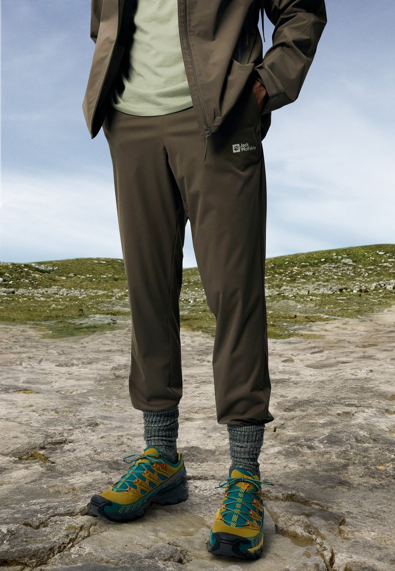 Vannavstøtende brune utendørsbukser med elastiske mansjetter, parret med fargerike turstøvler og teksturerte grå sokker. Jack Wolfskin-logo synlig.