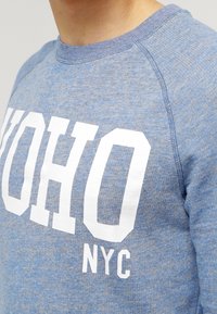 Blue heathered sweatshirt featuring bold white "UOHO NYC" text, round neckline, and ribbed cuffs for a textured finish.