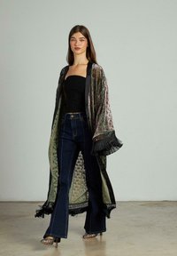 Kimono lung, cu un model complex, dintr-un material verde și negru, cu accente de franjuri, purtat peste un top negru și blugi înalți, de culoare închisă.