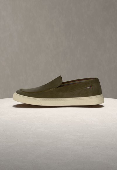 Alyvuogių žalios spalvos zomšinis batelis be raištelių su balta guminė padu, pastatytas ant šviesios dangos priešais vienspalvį foną.