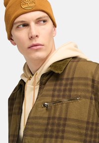 Brunternetternetternet jakke med ternet mønster og en lynlås lomme, båret over en beige hoodie med gule accenter, parret med en sennepsfarvet strikket hue.
