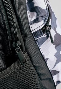 Sac à dos camouflage noir et gris avec des caractéristiques : matériau en nylon texturé, zippers bien visibles, poche latérale en mesh et motifs de couleur distincts.