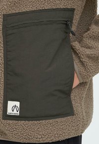 Giacca in pile color sabbia con tasca verde oliva, dotata di chiusura con zip e patch del logo. La texture è morbida e pelosa.