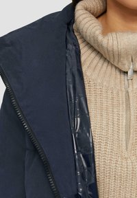 Marineblauer Daunenjacke mit glattem Innenfutter und strukturiertem, beigen Rollkragenpullover. Verfügt über einen Reißverschluss und einen gerippten Kragen.