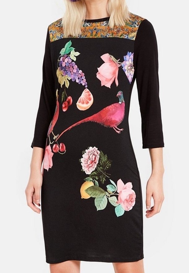 Robe noire à manches 3/4 avec des imprimés colorés de fleurs, fruits et oiseaux roses, portée par une personne aux cheveux blonds.