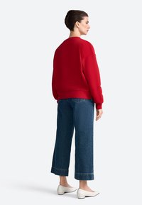 Červená mikina s okrúhlym výstrihom a rebrovanými manžetami, v kombinácii so širokými modrými džínsami a bielymi nízkymi topánkami na podpätku.