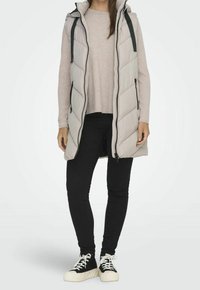 Gilet imbottito grigio chiaro con zip frontale, cappuccio e motivo a chevron, abbinato a un maglione beige e jeans skinny neri. Sneakers bianche e nere.