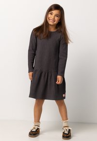 Lächelndes Mädchen mit langen braunen Haaren, das ein dunkelgraues langärmliges Kleid, cremefarbene Rüschensocken und Sneakers mit Leopardenmuster trägt und auf einem weißen Boden steht.