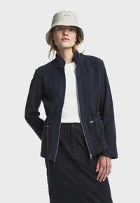 Chaqueta de forro polar azul marino con cuello alto, bolsillos con cremallera y cordones ajustables en la cintura; superpuesta sobre una blusa blanca.