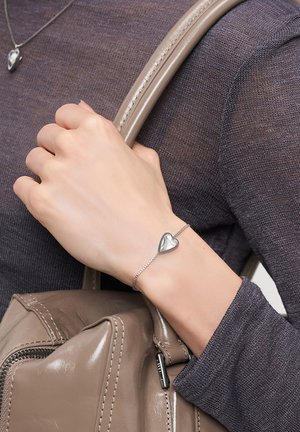 Hand wearing a silver heart-shaped bracelet holds beige leather handbag strap, wearing dark brown knit top.