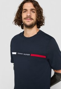 Marineblaue Baumwoll-T-Shirt mit einem horizontalen Streifen in Weiß und Rot und dem Text "TOMMY HILFIGER" in der Mitte.