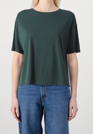 Personne portant un t-shirt ample à manches courtes vert foncé et un jean denim bleu sur un fond clair uni.