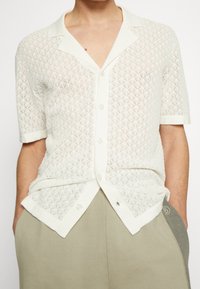 Camisa blanca de manga corta con un patrón de encaje en forma de diamante, que cuenta con un cuello suave y botones. Combinada con pantalones gris claro.