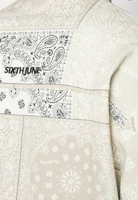 Tessuto beige stampato con design paisley e accenti neri. Presenta il logo "SIXTHJUNE", texture liscia e cuciture a contrasto.