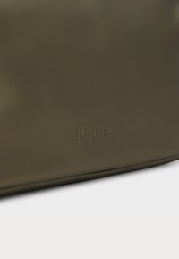 Tamsiai alyvuogių spalvos, vandeniui atspari kuprinė su glotnia tekstūra ir iškilniu "RAINS" logotipu. Pasižymi minimalistišku dizainu ir elegantiška stačiakampio forma.