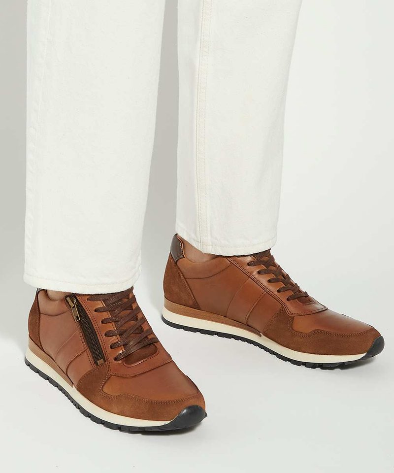 Baskets en cuir marron et daim avec zip latéral et lacets portées avec un pantalon blanc sur un fond clair.