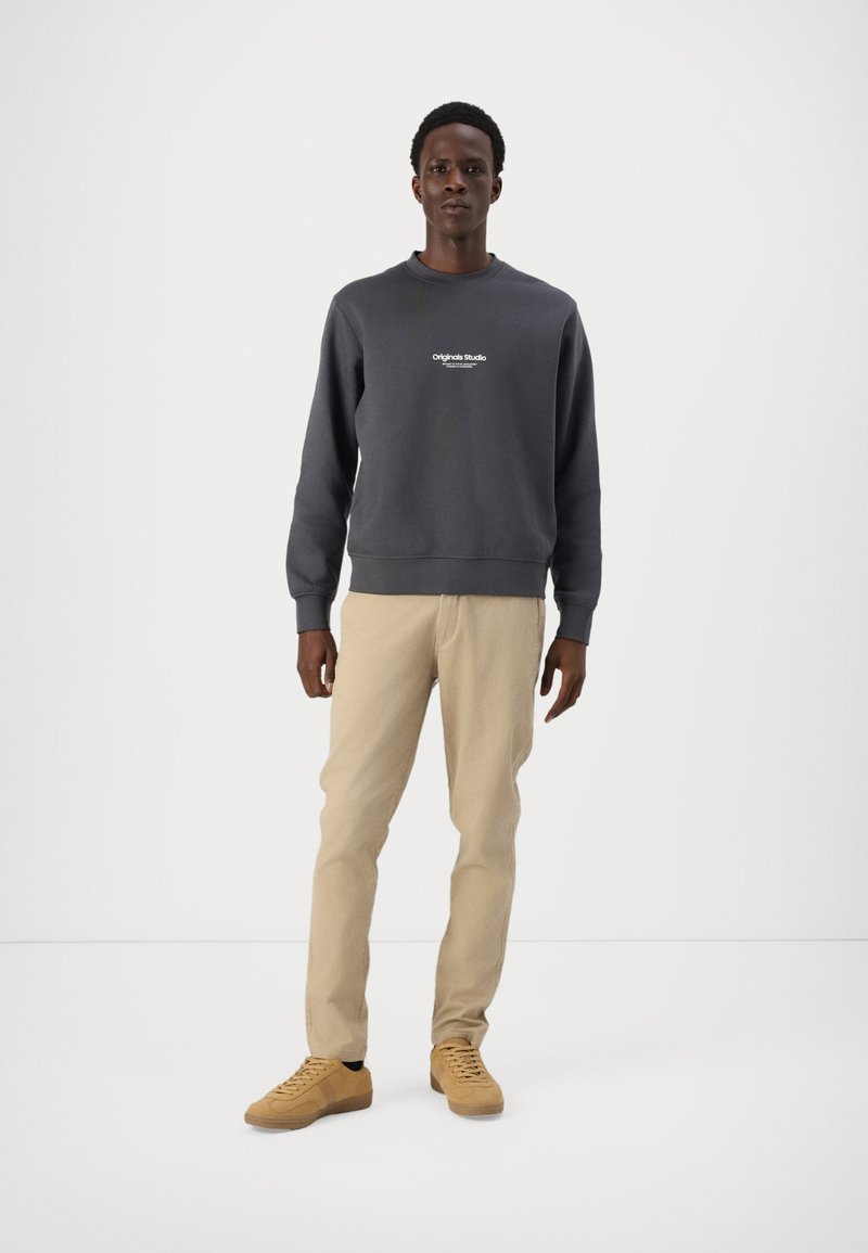 Felpa grigio scuro a girocollo con dettaglio del logo bianco, abbinata a pantaloni beige chiaro e sneakers beige con finitura texturizzata.