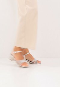 Stříbrné sandály s pásky a otevřenou špičkou, zdobené třpytivými detaily a nízkým blokovým podpatkem. Hladký povrch a nastavitelný pásek kolem kotníku.