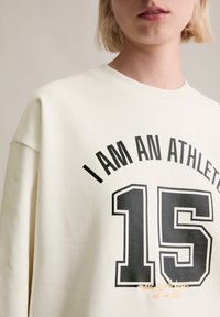 Osoba w kremowej bluzie z czarnym napisem "JESTEM SPORTOWCEM" nad dużą cyfrą "15" z przodu, na neutralnym tle.