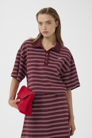 Ung kvinde med vådt hår iført en polo med bordeaux og hvide striber og nederdel, der holder en rød clutch.