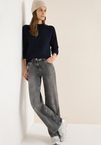 Maglione blu navy a coste, jeans grigi a gamba larga con effetto sbiadito e scarpe sportive argentate. Il modello indossa un berretto lavorato a maglia beige chiaro.