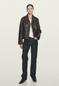 Donna che indossa una giacca di pelle marrone scuro, una camicia bianca, jeans neri a gamba dritta e scarpe nere a punta, in piedi con una mano in tasca.