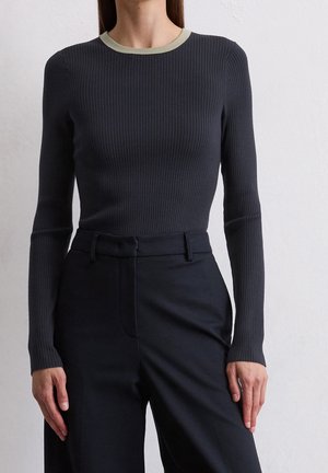 Kvinde iført en tætsiddende, ribstrikket sort langærmet top med en lys kontrast ved halsudskæringen og højtaljede sorte bukser mod en ensfarvet baggrund.