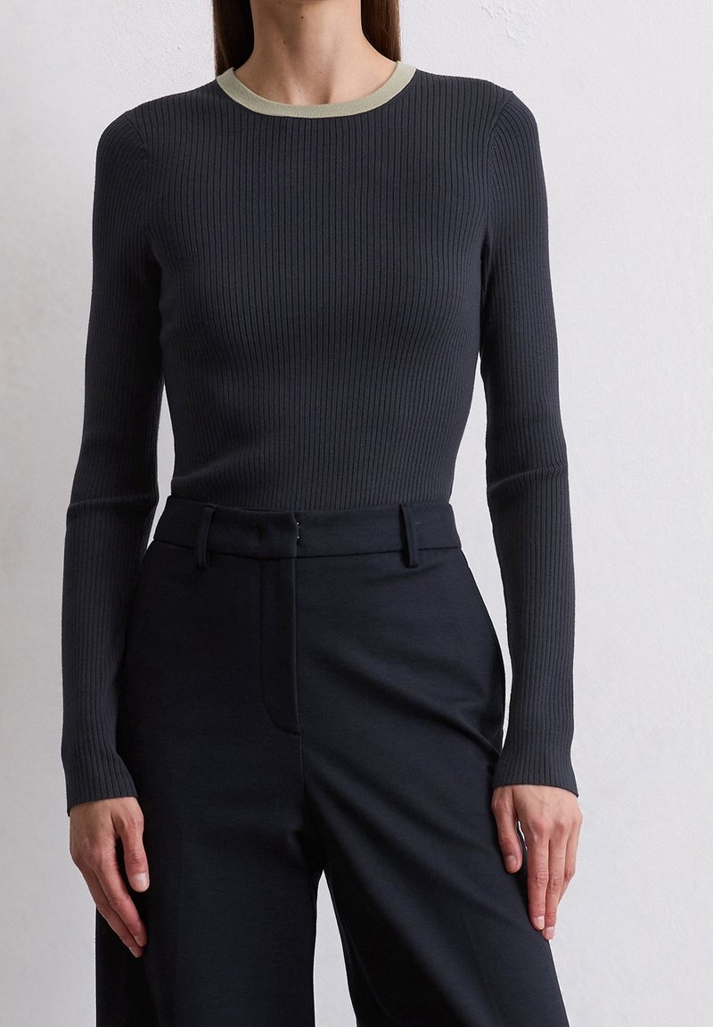 Femme portant un haut noir ajusté à manches longues côtelées avec un col clair contrastant et un pantalon noir taille haute, sur un fond uni.