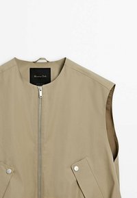 Gilet beige sans manches avec fermeture éclair à l'avant, col rond, deux poches à pressions et une boucle pour suspendre au col.