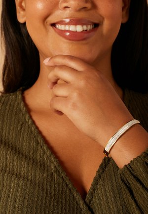 Gold-tone bracelet with a clear rhinestone accent. Wearer's hand resting on chin, wearing an olive green textured top. Smooth skin texture.