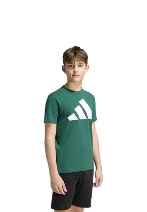 Un garçon porte un tee-shirt vert orné d’un grand logo Adidas blanc et un short noir sur fond blanc.