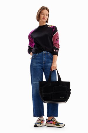 Borsa a tracolla nera con multiple tasche frontali, materiale in canvas e logo. Il modello indossa un maglione nero con maniche a fantasia colorata e jeans blu.