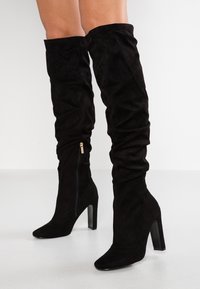 Botas de ante negras hasta la rodilla con un diseño arrugado, punta afilada, tacón cuadrado alto y cierre de cremallera lateral. Textura suave en toda su superficie.