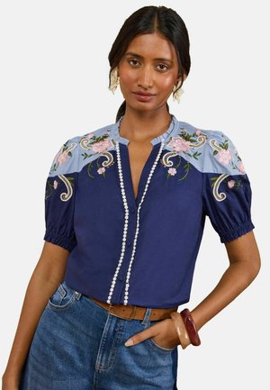 Frau trägt eine marineblaue Bluse mit hellblauem Passe, rosa Blumenstickerei, kurzen Puffärmeln, braunem Gürtel, Jeans und klobigen Armbändern.
